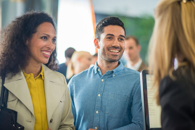 a smiling couple talking to a credit union representative with people in line behind them.