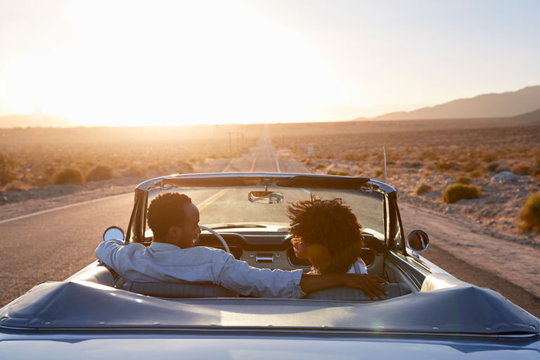 A couple in a convertible driving down a desert road.