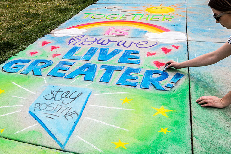 Woman drawing a chalk drawing on a sidewalk. The drawing says "Together is how we Live Greater!" With a smiling sun, rainbow, and stars. Also, there is a blue Nevada that says "Stay Positive." 