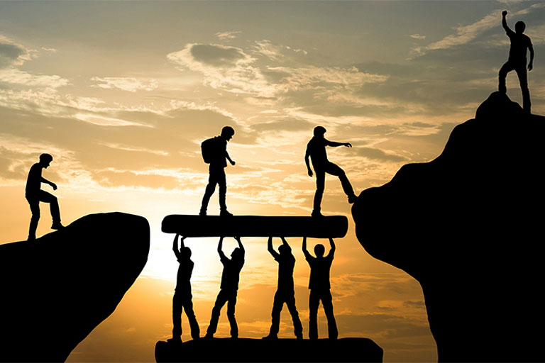 Silhouettes of people holding up a log to help others cross a gap in a cliff. 