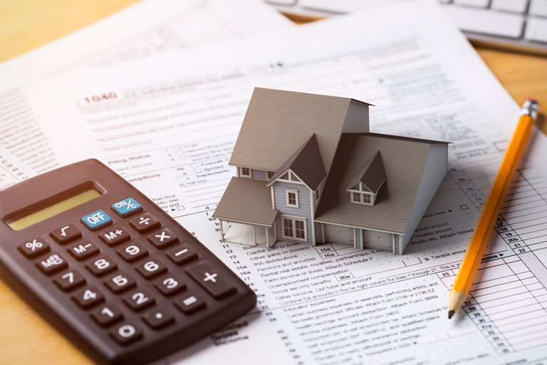 A calculator next to a tiny model house and a pencil on top of tax forms.