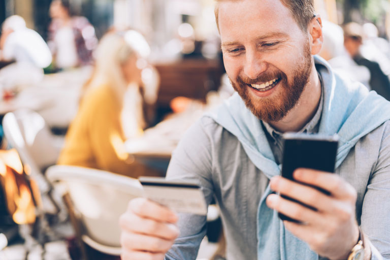 A man with a sweater tied around his neck holding a smartphone in one hand and a credit card on the other at a restaurant or café. 