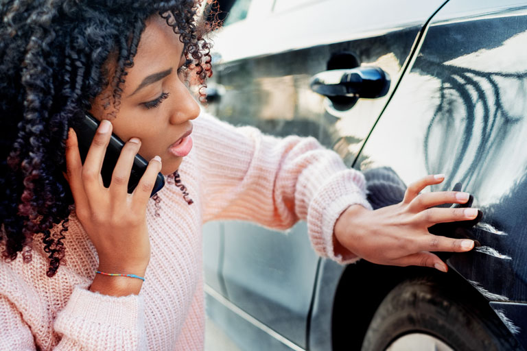 A woman on the phone assessing damage to her car by the left rear wheel well.