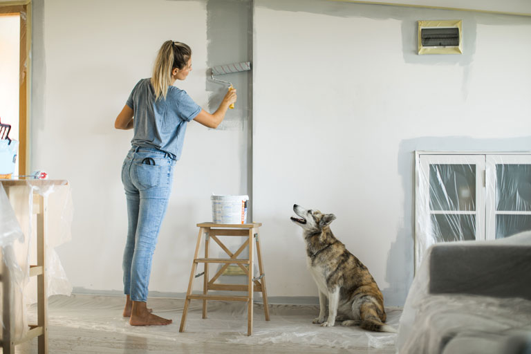 A woman painting a new house with her dog watching her. The dog is cute.