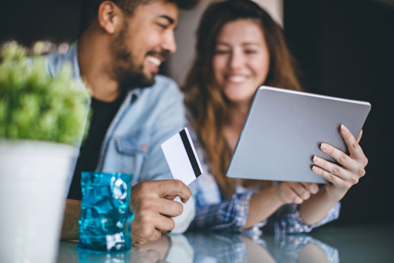 Couple sitting at a table with the man holding a credit card and the woman holding a tablet.