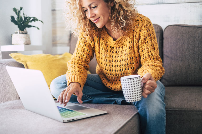 Woman holding a cup of coffee on her laptop sitting on a couch.