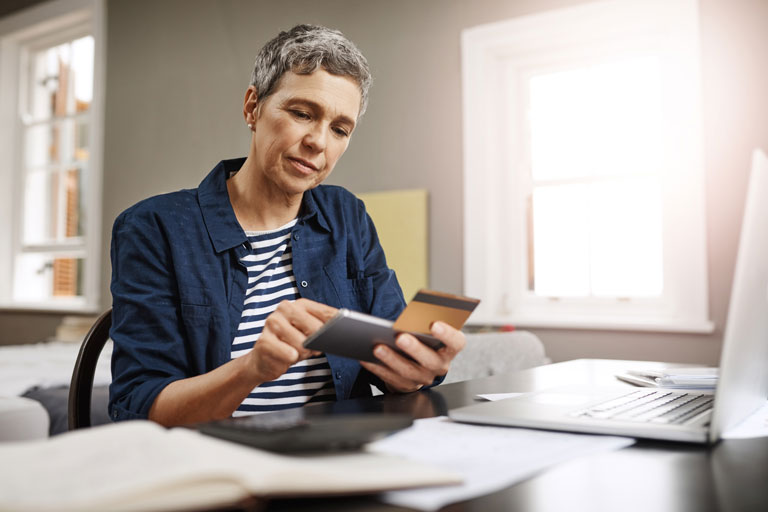 Woman in her late 50s with a smart phone and credit card in hand sitting at a table with a laptop. 