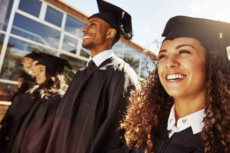 A line of young students dressed in graduation gowns and caps wait outside to graduate.