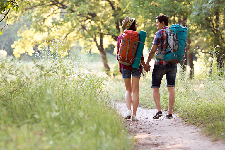 Sporty Family Man and Woman with Backpacks walking along Forest Trail holding Hands with Sunny Background