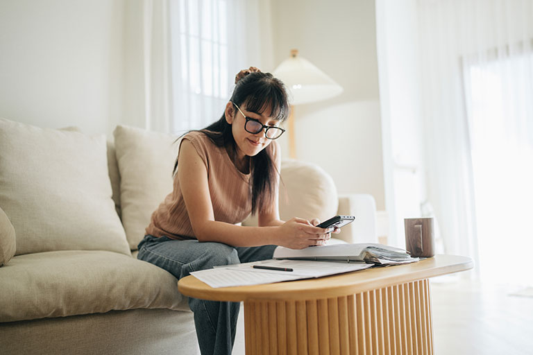 A woman sits at her living room with smartphone and financial reports doing her monthly budget.