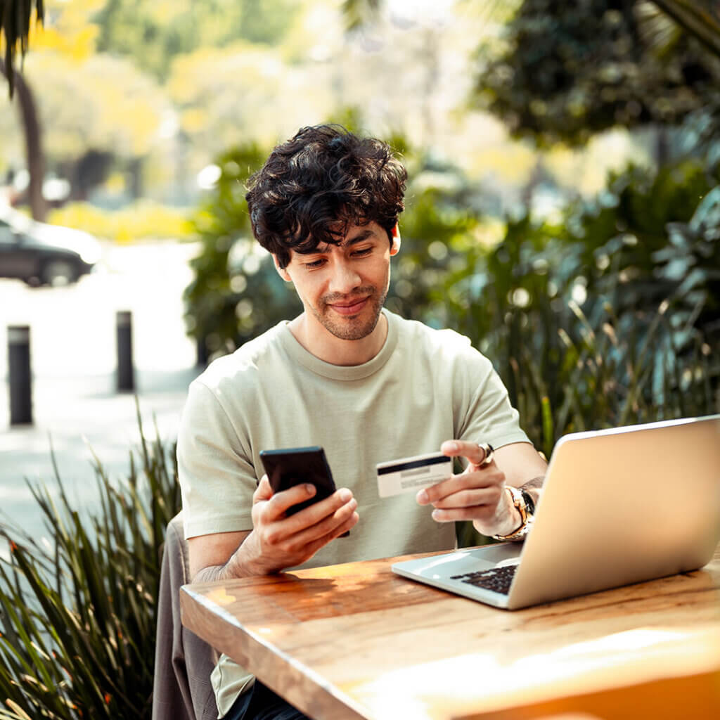 Young man sitting outside looking at his phone, card, and laptop