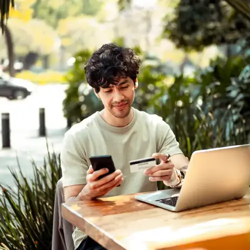 Young man sitting outside looking at his phone, card, and laptop
