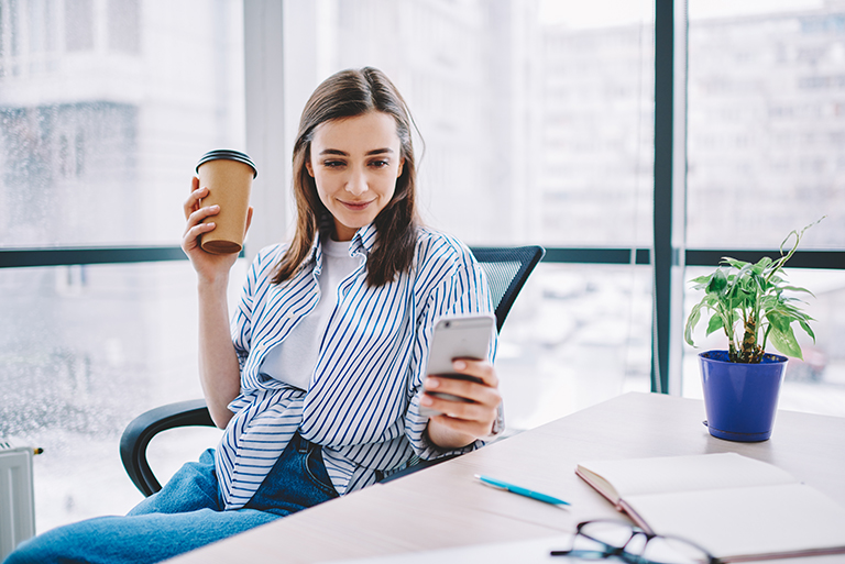 Young woman looking at her phone to learn about  the different essential bank accounts while sipping coffee